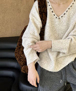 Vネックの縁に黒いステッチが入ったアイボリーのオーバーサイズニットを着用し、ソファーに座る女性