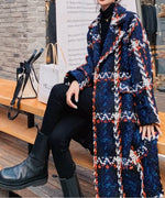 大柄チェックのツイードロングコートを羽織り、黒のタートルネックと黒のロングブーツを合わせた女性が足を組んでいる写真