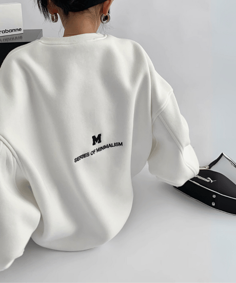 白いオーバーサイズスウェットを着用して座る女性の背中側。小さな「M」ロゴと「SERIES OF MINIMALISM」の英文プリントが施されている。