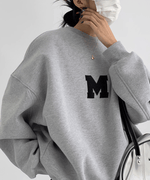 グレーのオーバーサイズスウェットの上半身クローズアップ。胸元の黒い「M」ロゴと、ドロップショルダーの切り替え線が見える。