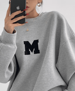 グレーのオーバーサイズスウェットの上半身クローズアップ。胸元の黒い「M」ロゴが強調されたショット。