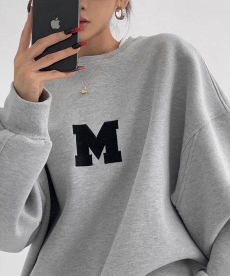 グレーのオーバーサイズスウェットの上半身クローズアップ。胸元の黒い「M」ロゴが強調されたショット。