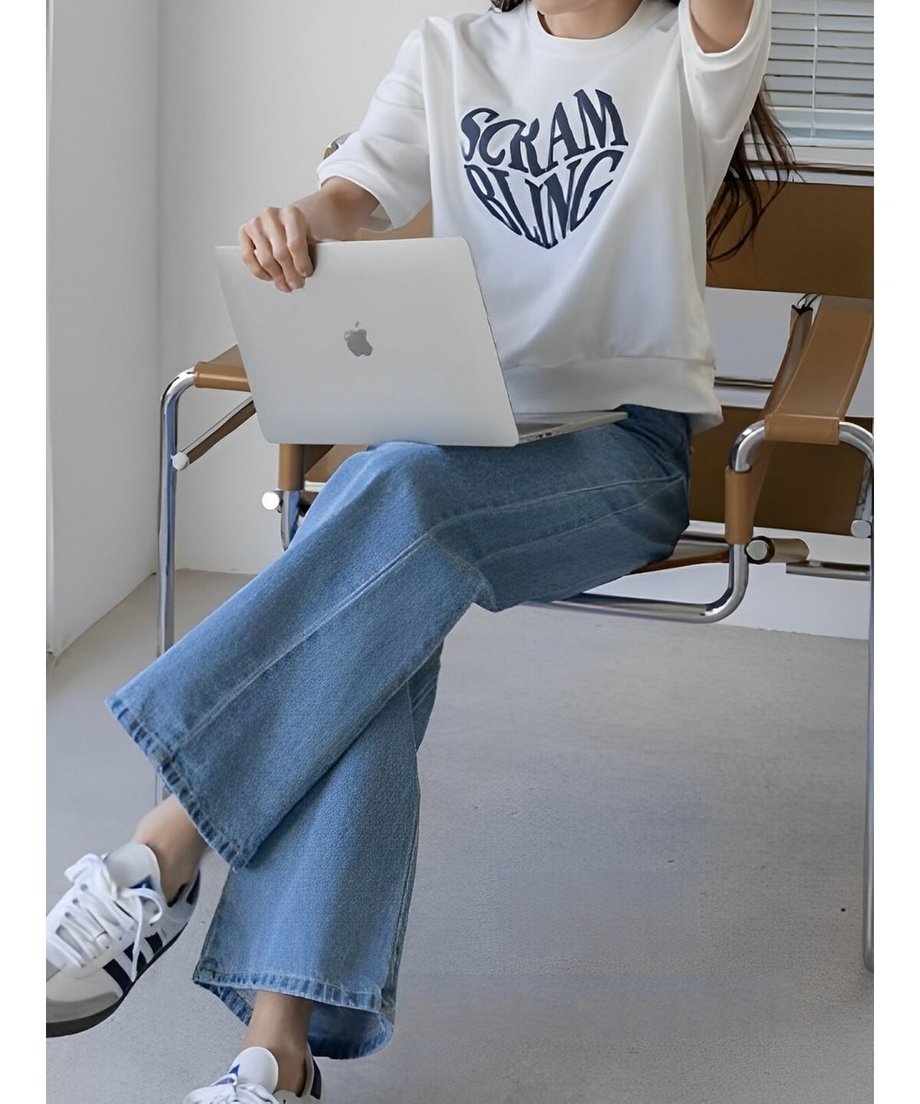 ホワイトのTシャツを着た女性が、青いデニムパンツを履き、椅子に座ってノートパソコンを操作している様子。