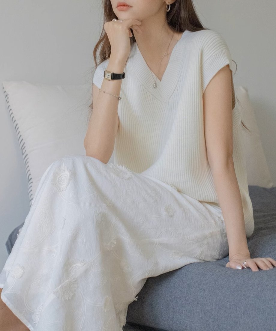 ホワイトのニットベストにロングスカートを合わせた女性がソファーに座っている。
