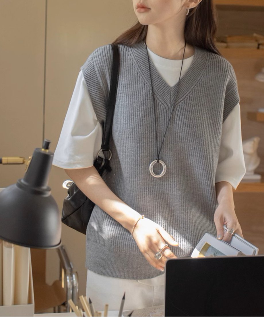 グレーのニットベストを着た女性が肩にバックをかけて立っている姿。