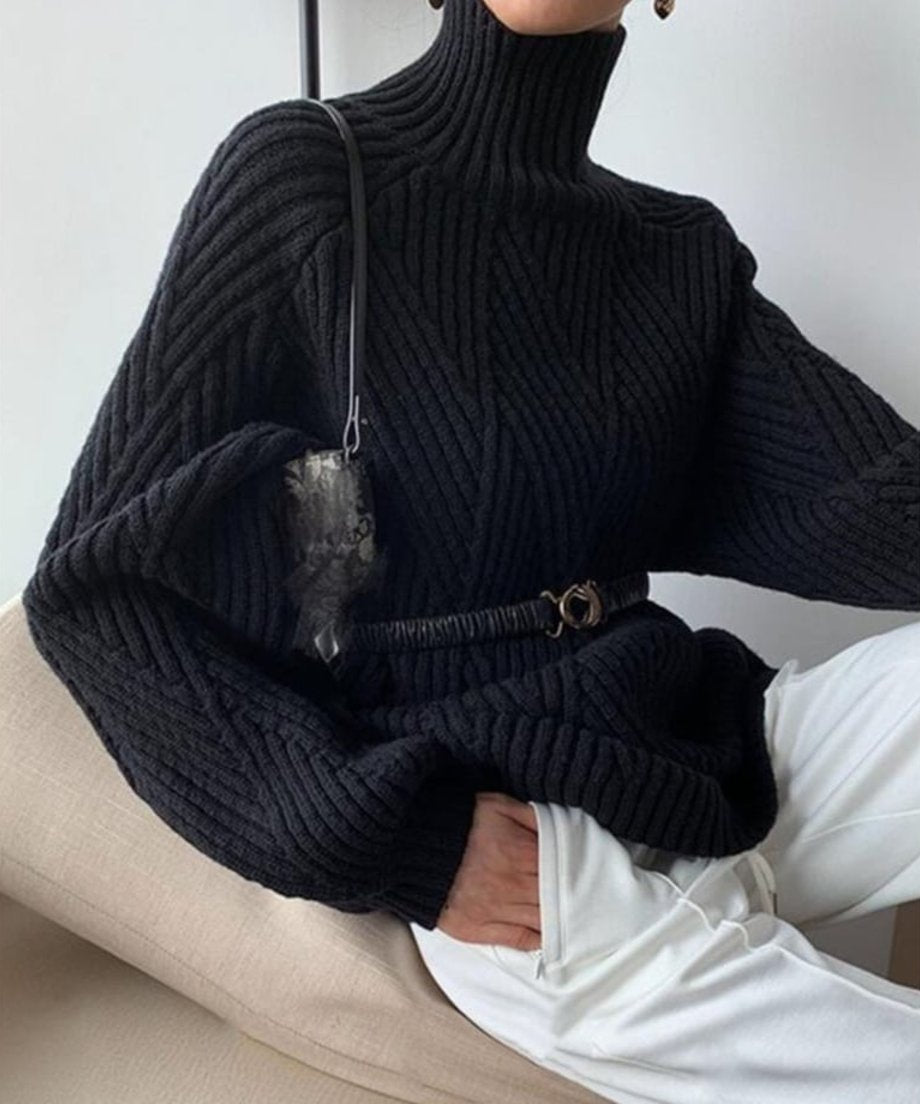 ブラックの立体編みタートルネックセーターとホワイトパンツを着用した女性がソファーに座っている