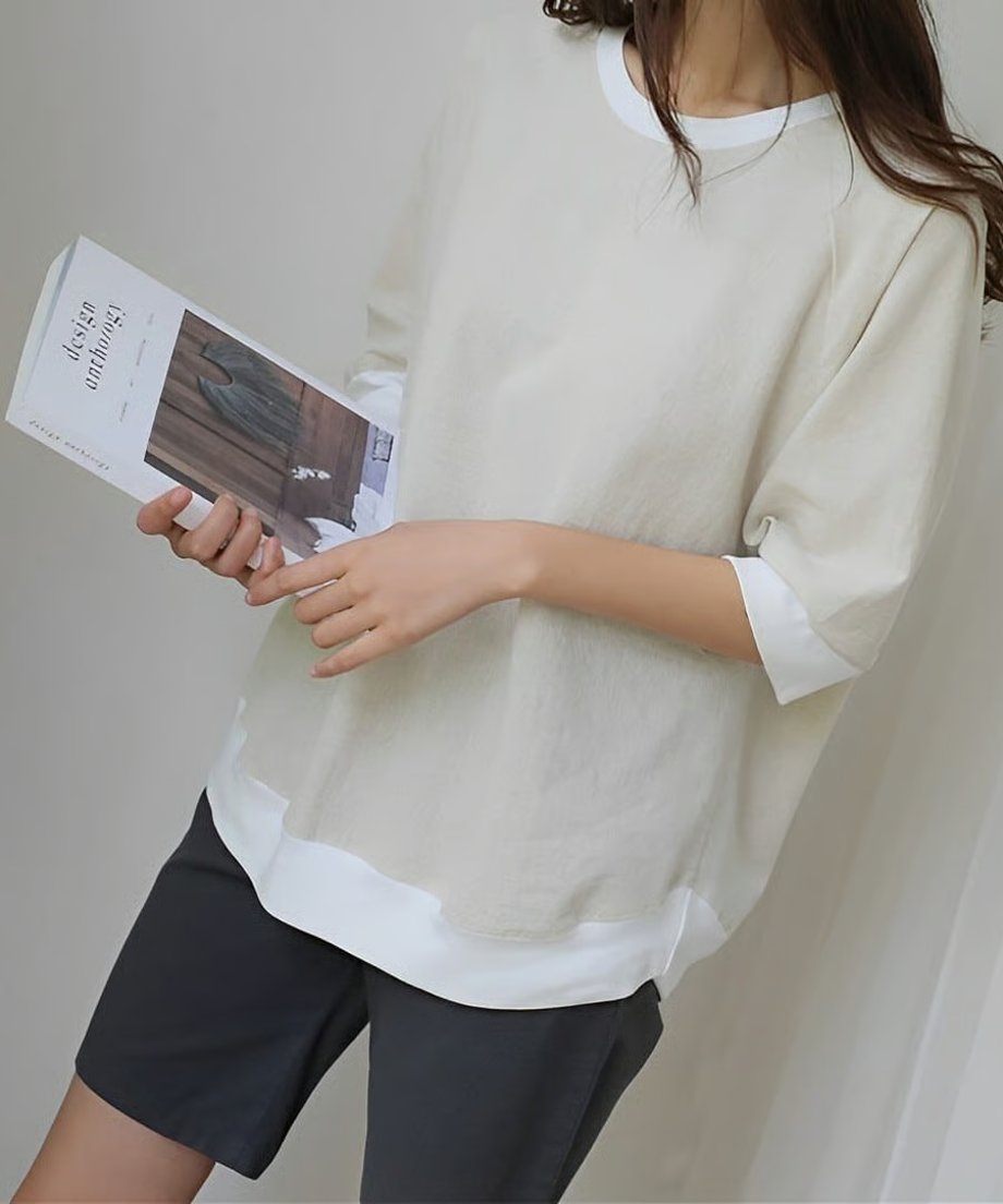 アイボリーのカジュアルトップスを着用した女性が手に本を持つ画像。