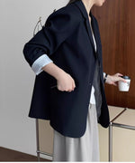グレーのパンツと白いトップスにネイビーのブレザーを羽織った女性。ポケットに手を入れている。