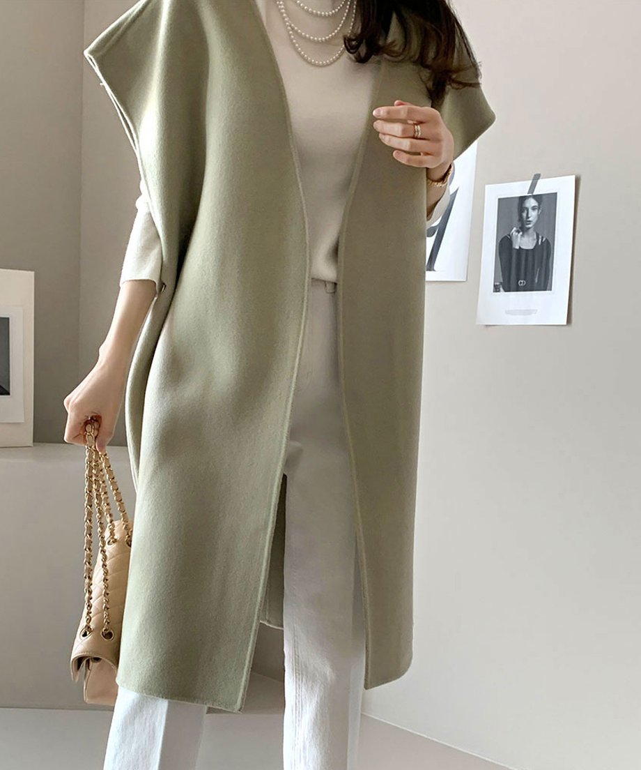 ライトグリーンのノースリーブコートを着た女性、ベージュのバッグとアクセサリーを身に着けて立っている写真