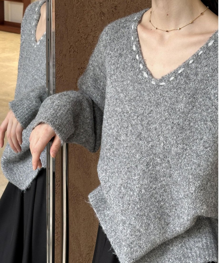 Vネックの縁に白いステッチが入ったグレーのオーバーサイズニットを着用した女性が鏡の横に立っている写真