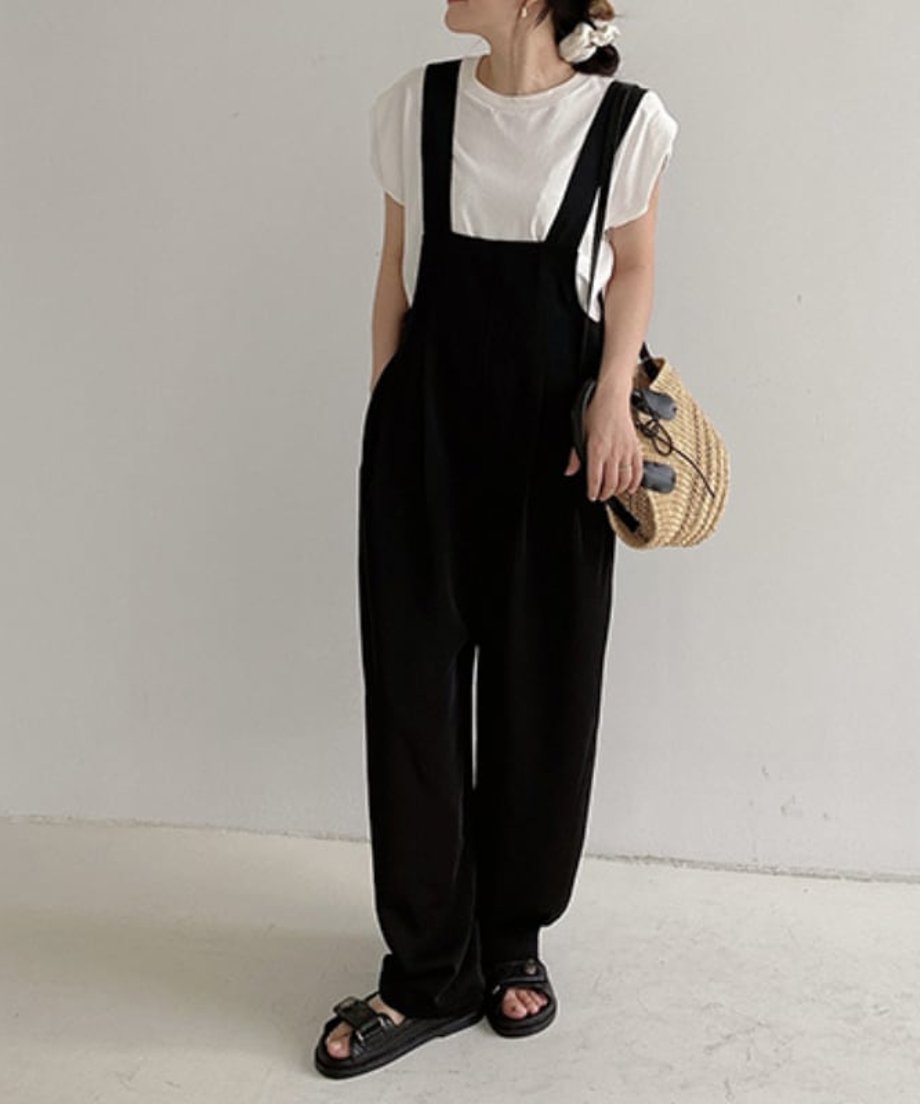 白いTシャツと黒いワイドレッグサロペットを着用し、ストロー素材のバッグを持った女性の全身。黒い厚底サンダルを履いている。