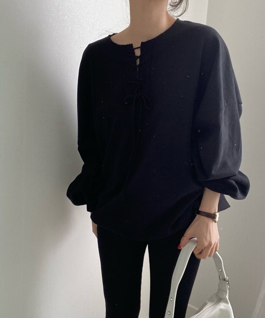 ブラックのレースアップネックプルオーバーを着用した女性の全身。黒のボトムスに白のショルダーバッグを持ち、シンプルな腕時計とリングを合わせたモノトーンコーデ。