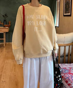 クリームのオーバーサイズスウェットと白のワイドパンツを着用した女性の全身。屋内での撮影で、スウェットのフロントロゴが見える。
