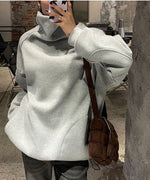グレーのタートルネックスウェットを着用し、キルティングバッグを斜めがけした女性のバストアップ。室内でスマートフォンを触っている。