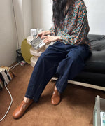 花柄のブラウスと濃紺のワイドデニムパンツ、ブラウンのレザーストラップシューズを着用した女性が、黒いソファに座り、本を持っている全身コーディネート画像。