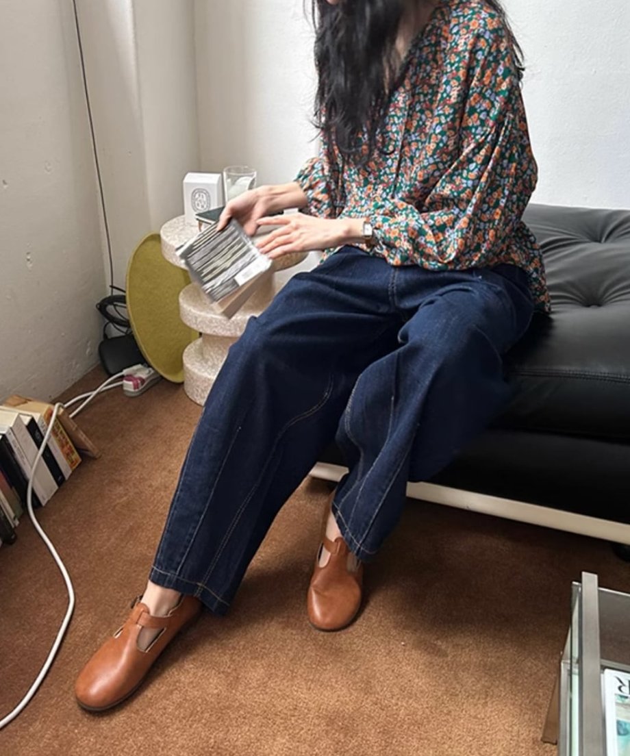 花柄のブラウスと濃紺のワイドデニムパンツ、ブラウンのレザーストラップシューズを着用した女性が、黒いソファに座り、本を持っている全身コーディネート画像。