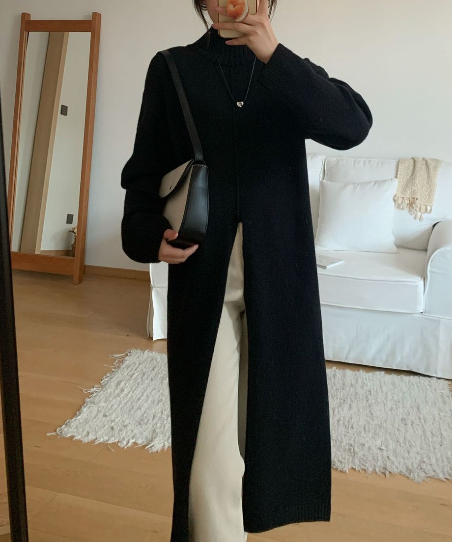 黒のタートルネックロングニットを着用した女性が、白いパンツとハイカットスニーカーを合わせ、木製の家具にもたれかかっている全身コーディネート写真。