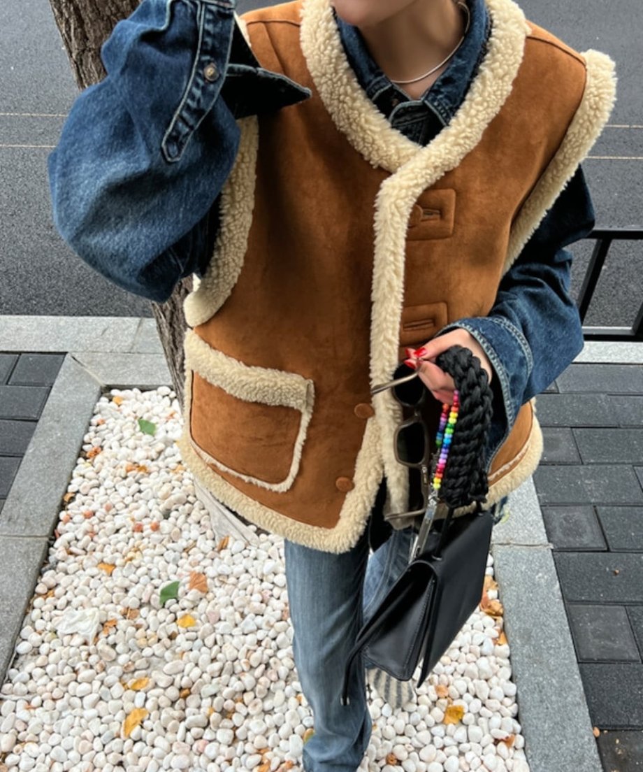 ブラウンとアイボリーのムートン風ベストをデニムジャケットとレイヤードし、濃淡デニムパンツと合わせたカジュアルな全身コーディネート。黒のショルダーバッグとカラフルなビーズのスマホストラップを手に持っている。