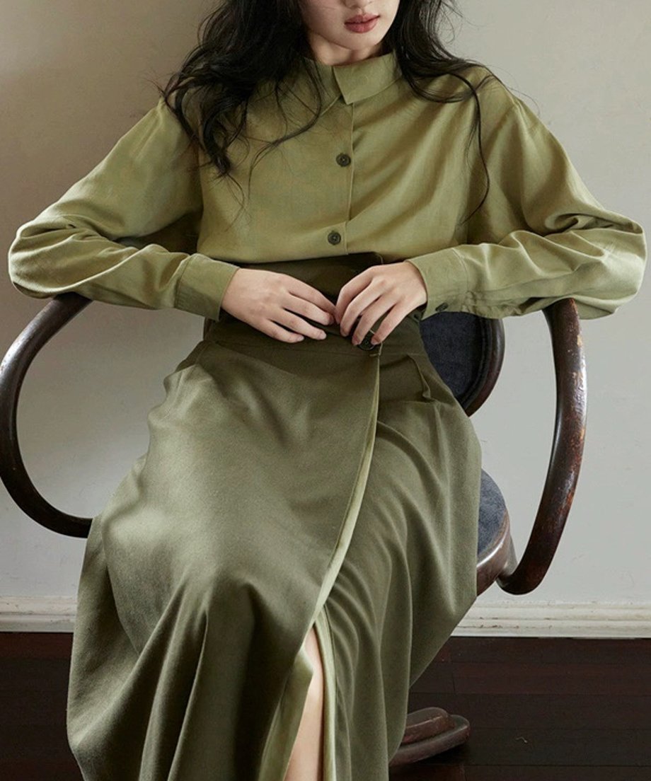 オリーブグリーンのセットアップを着た女性が木製の椅子に座っているショット。落ち着いたトーンのファッションポートレート。