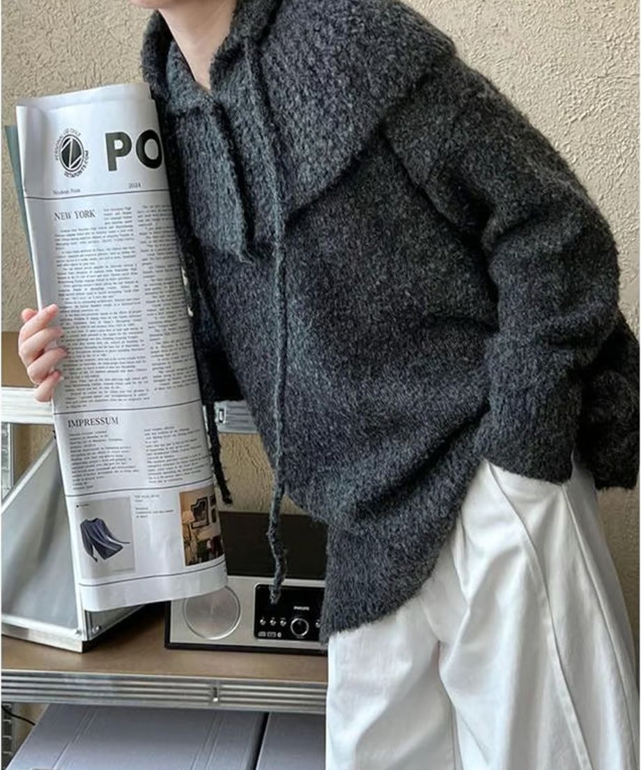 グレーのざっくり編みフード付きニットを着用した女性の全身。ゆったりとした白のパンツを合わせ、新聞を持ったカジュアルなスタイリング。