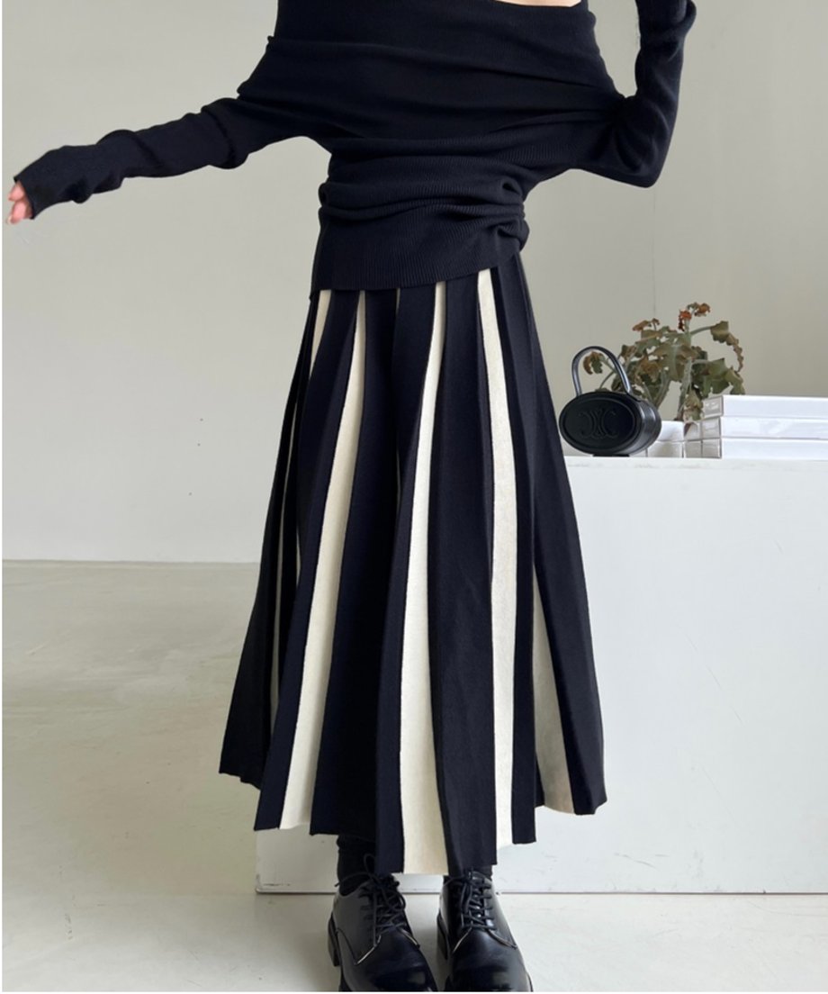 黒のリブニットと、黒とホワイトの太い縦ストライプが入ったロング丈のプリーツスカートを着用し、足元は黒いレースアップブーツ。