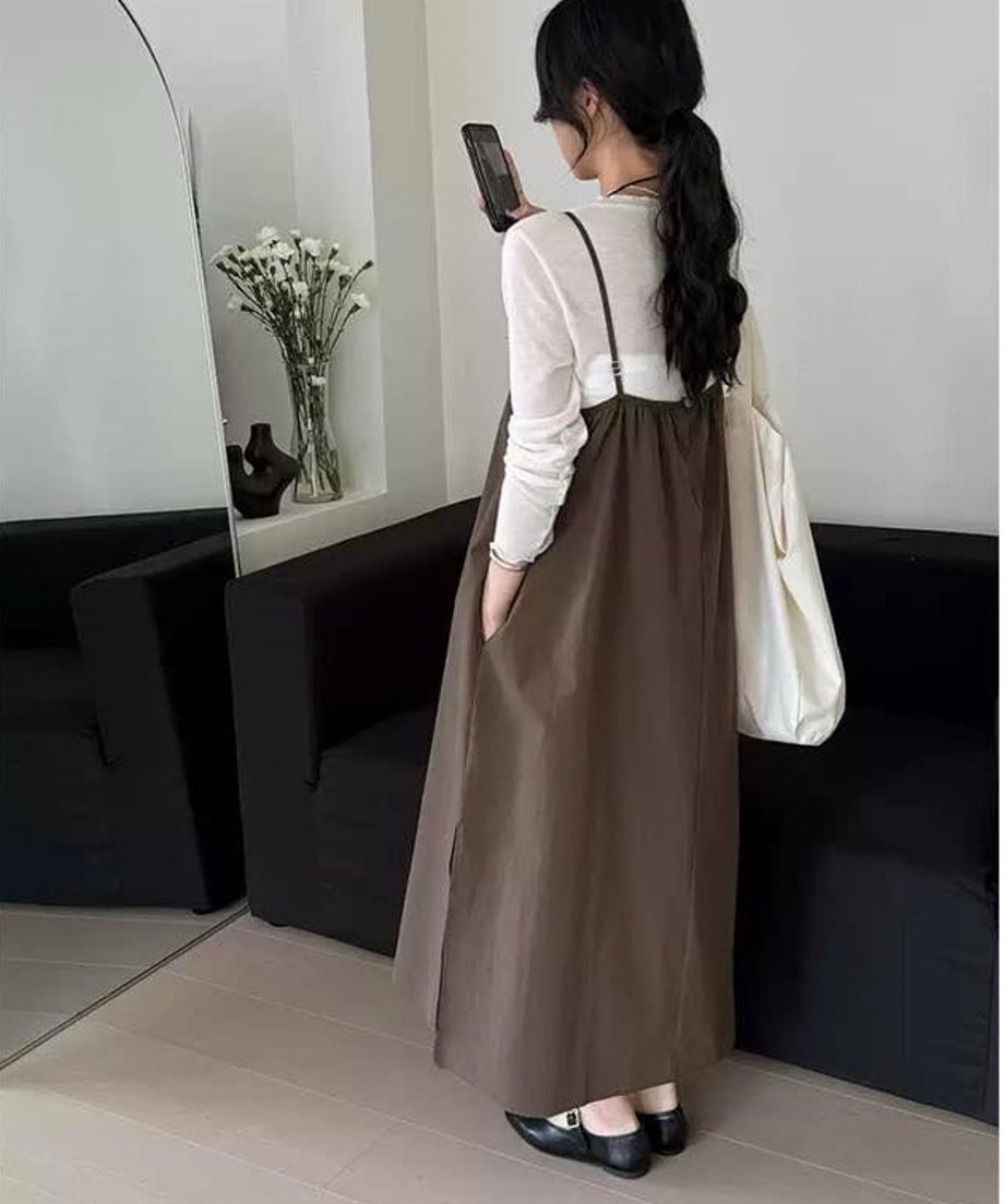 白い長袖トップスに茶色いロングキャミソールワンピースを着用し、スマートフォンを持っている女性のバックスタイル。