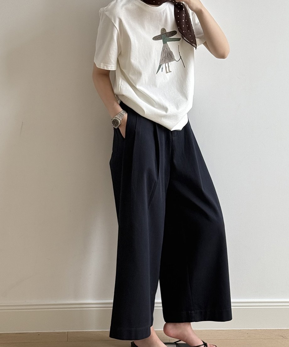 ネイビーのワイドパンツと、イラストがプリントされた白いTシャツを着用した女性の半身。手をポケットに入れ、スカーフを首元に巻いている。