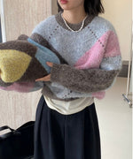 グレー/ピンクのアーガイル柄ニットを着用した女性のバストアップ。もう一つのブラウン系ニットを手に持ち、黒いボトムスと白インナー、パールネックレスを着用している。