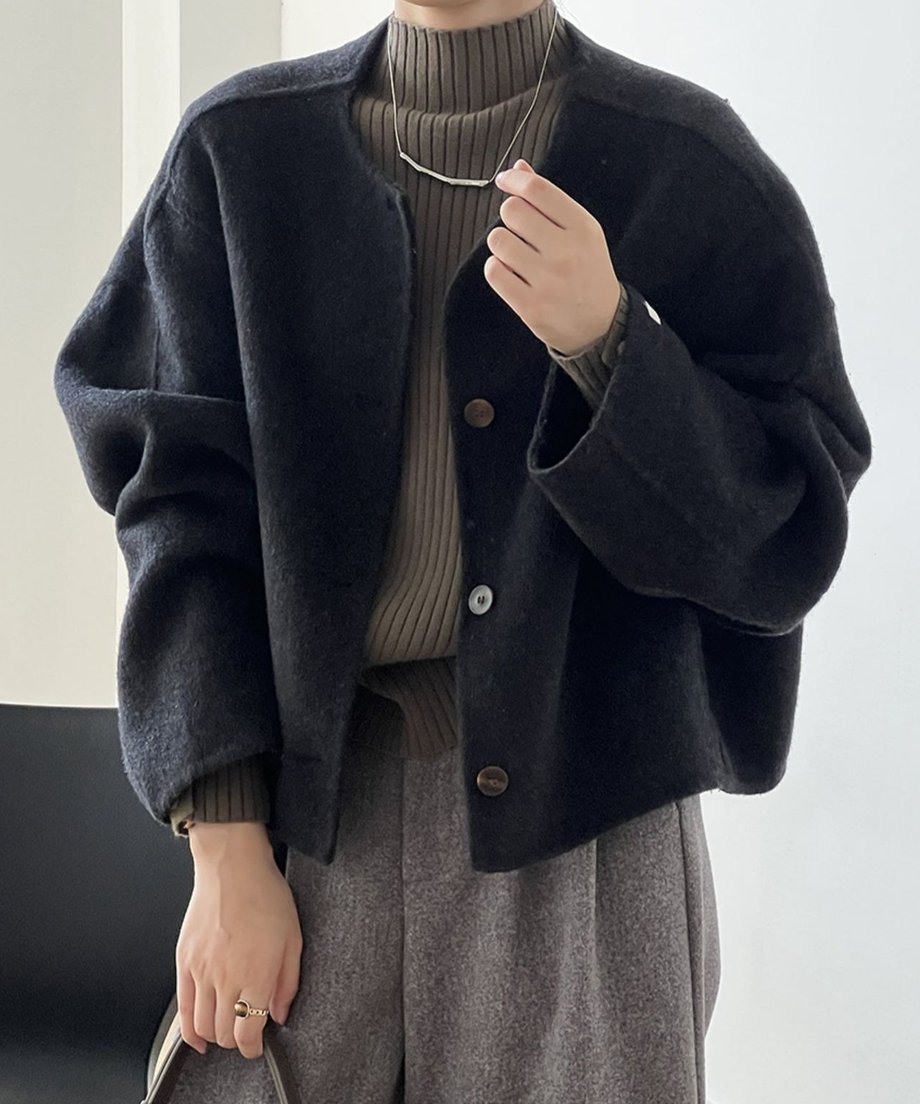 ダークグレーのカラーレスショートコートを着用した女性の正面。カーキ色のリブタートルネックと茶色のチェック柄パンツを合わせ、ネックレスを着用している。