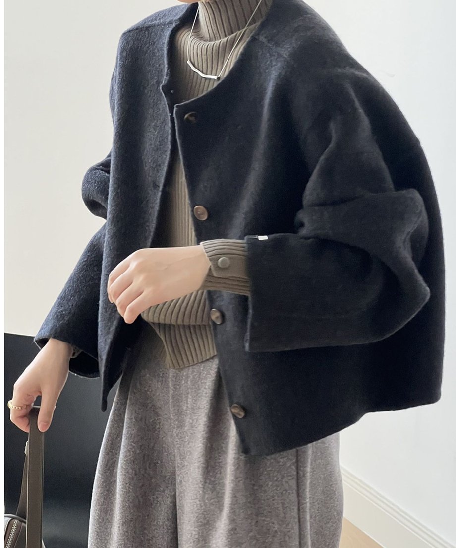 ダークグレーのカラーレスショートコートを着用した女性の側面。カーキ色のハイネックリブニットとグレーのボトムスを合わせ、大きなバッグを持っている。