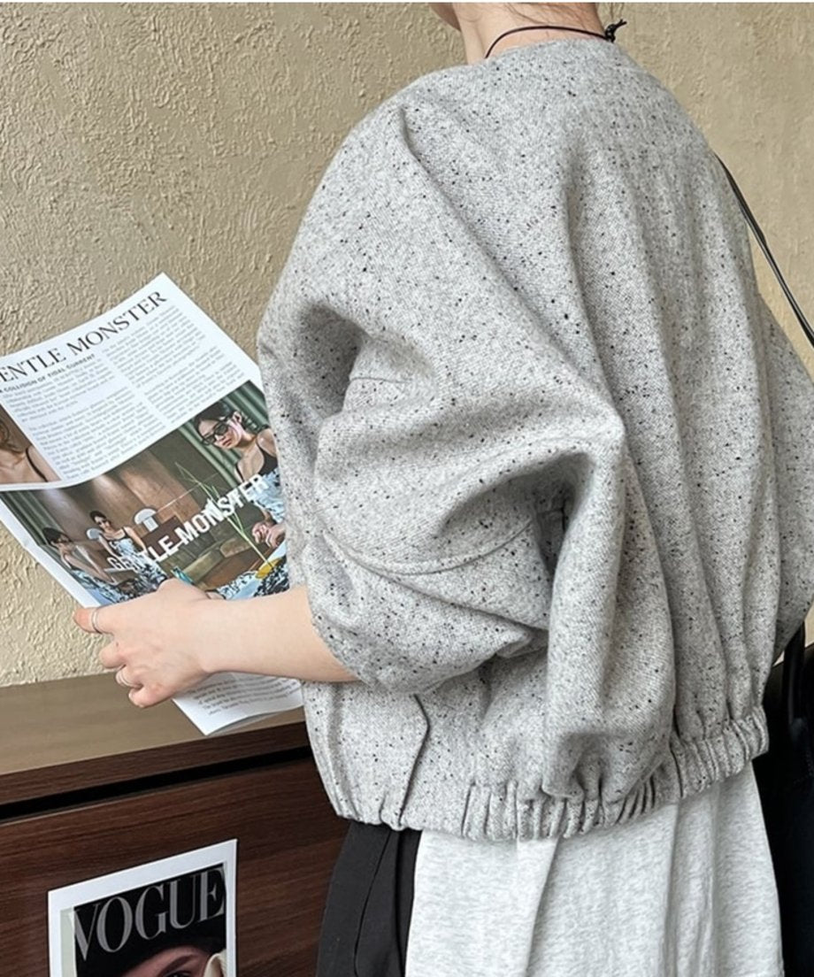 グレーのデザインジャケットを着用し、背面から見たクローズアップ。手には雑誌を持ち、裾のシャーリングによるギャザーと丸みのあるバックスタイルが見える。