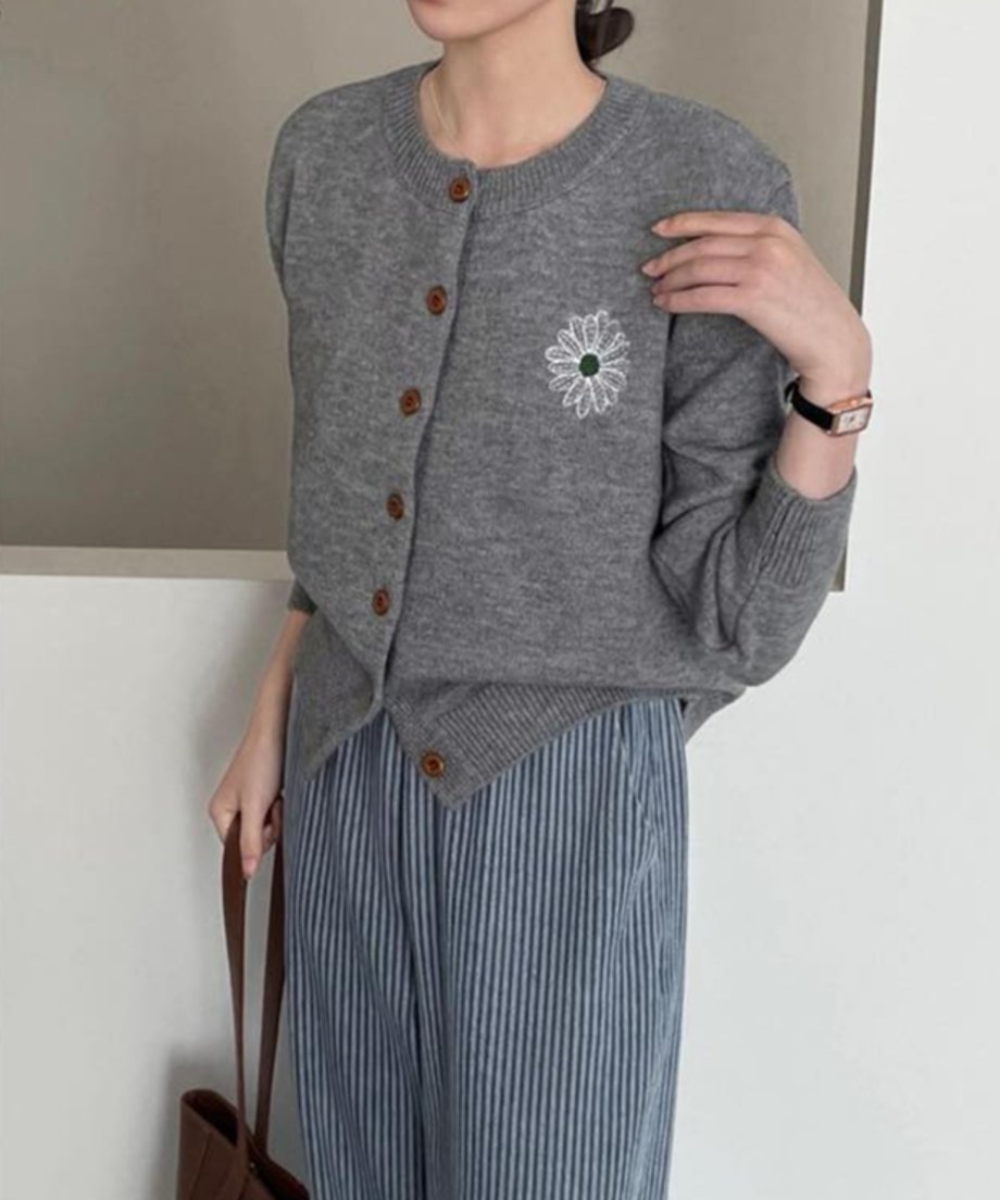グレーの花柄刺繡ニットカーディガンを着用した女性の半身像。白い花と緑の中心を持つ刺繍が左胸にあり、青と白のストライプ柄のパンツと茶色のトートバッグを合わせている。