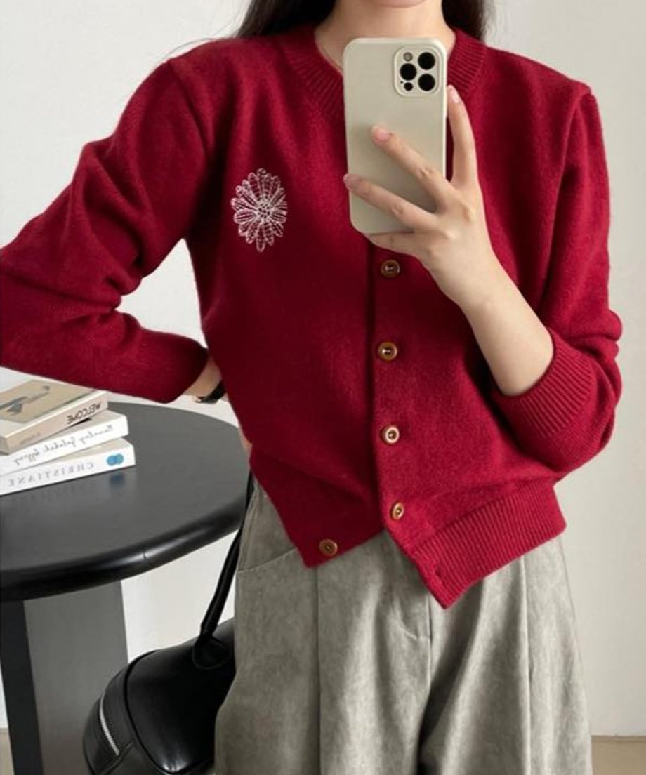 レッドの花柄刺繡ニットカーディガンを着用した女性のクローズアップ。グレーのワイドパンツを合わせ、黒いテーブルの上に置かれた本が背景に見える。