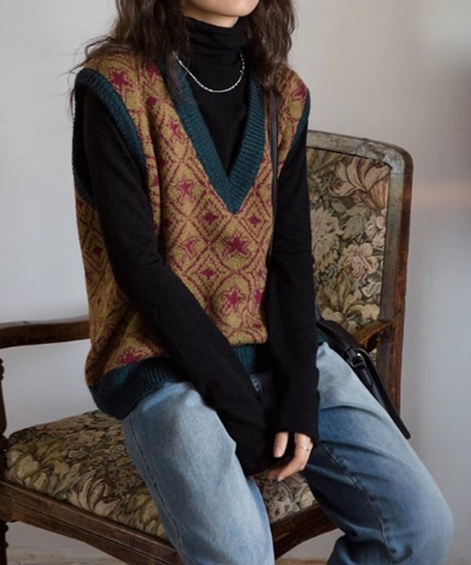 レトロ調Vネックニットベストを着用し、椅子に座っている女性の正面ショット。シルバーのネックレスを着用し、タートルネックとベストのレイヤードが見える角度です。