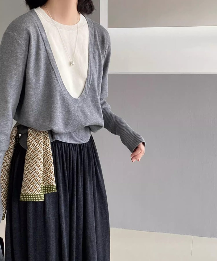 グレーのディープUネックニットを着用した女性の半身像。白いTシャツをインナーに重ね、黒いロングスカートを着用し、柄のスカーフを腰に巻いている。