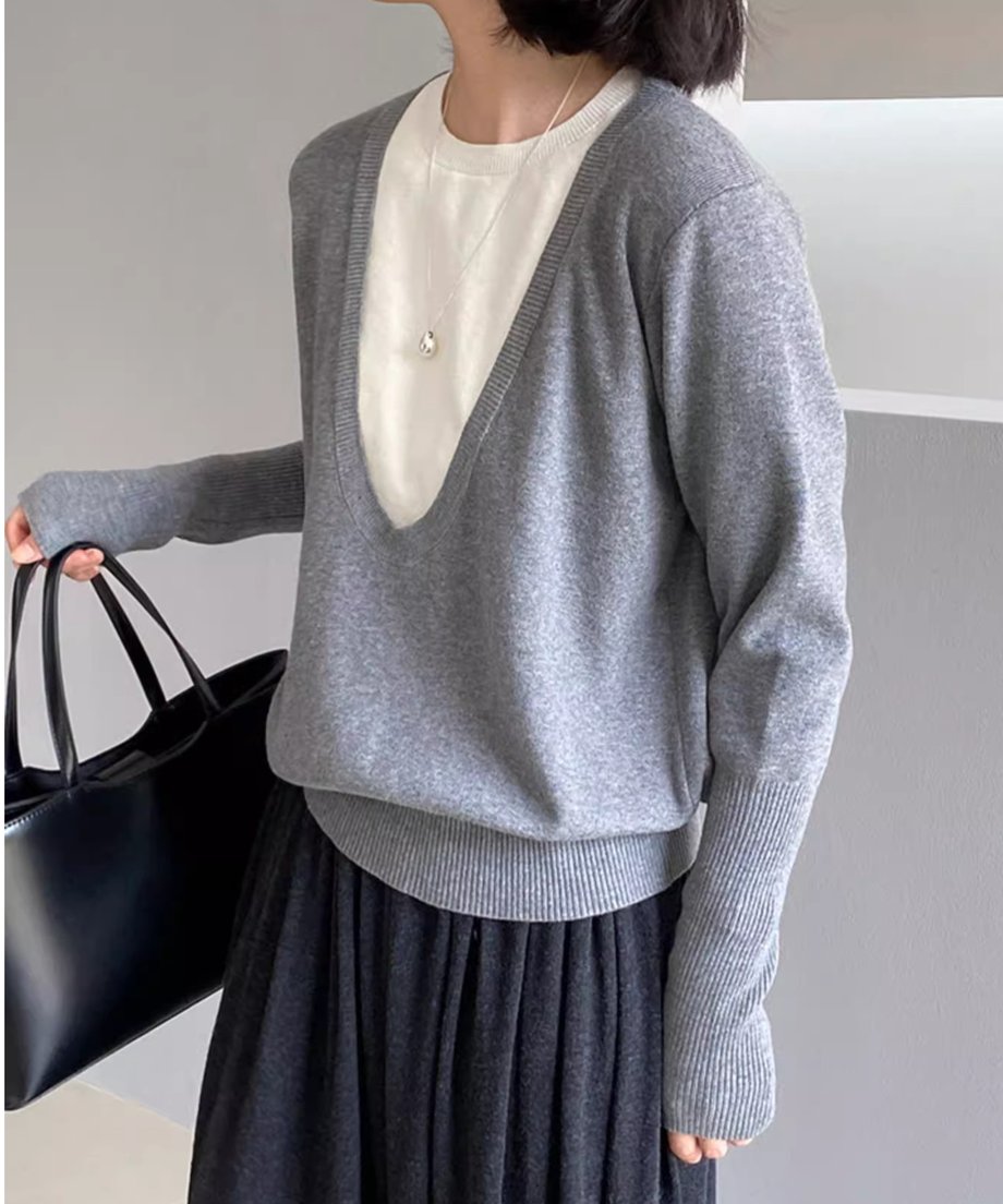 グレーのディープUネックニットを着用した女性の半身像。白いTシャツと黒いプリーツスカートを着用し、黒いトートバッグを手に持っている。