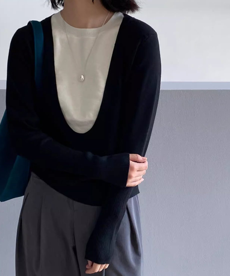 ブラックのディープUネックニットを着用した女性の半身像。白いTシャツをインナーに重ね、グレーのワイドパンツを着用。