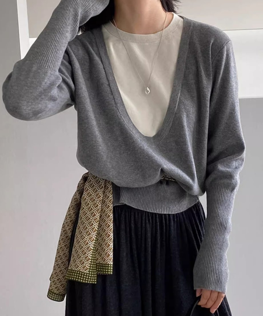 グレーのディープUネックニットを着用した女性の半身像。白いTシャツと黒いロングスカートを着用し、柄のスカーフを腰に巻いている。