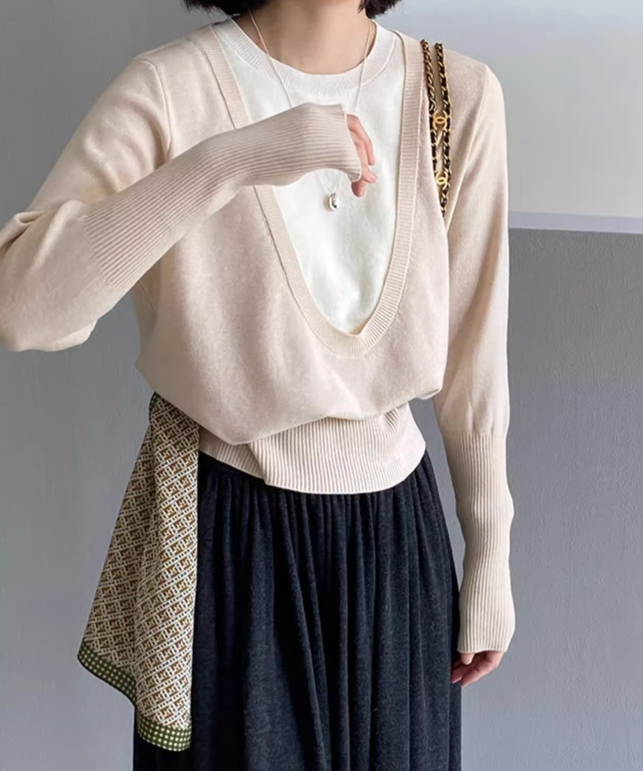 アプリコットのディープUネックニットを着用した女性の半身像。白いTシャツをインナーに重ね、黒いスカートを着用し、柄のスカーフを腰に巻いている。