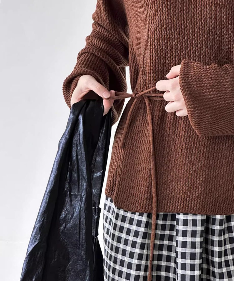 ブラウンのホールデザインニットを着用した女性が、同素材の細いウエストリボンを背中の部分で結んでいる手のクローズアップ。