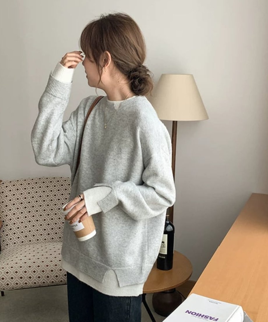 ライトグレーのオーバーサイズクルーネックニットを着用した女性が、横向きで立ち、片手にカップを持っている。裾と袖口のディテールが見えるレイヤード風のカジュアルな着こなし。