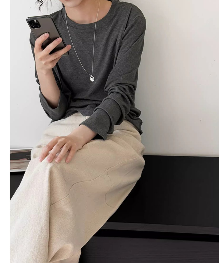 アイボリーのワイドパンツ（スカート状に見える）とチャコールグレーの長袖Tシャツを着て携帯電話を見ている女性が座っている写真。