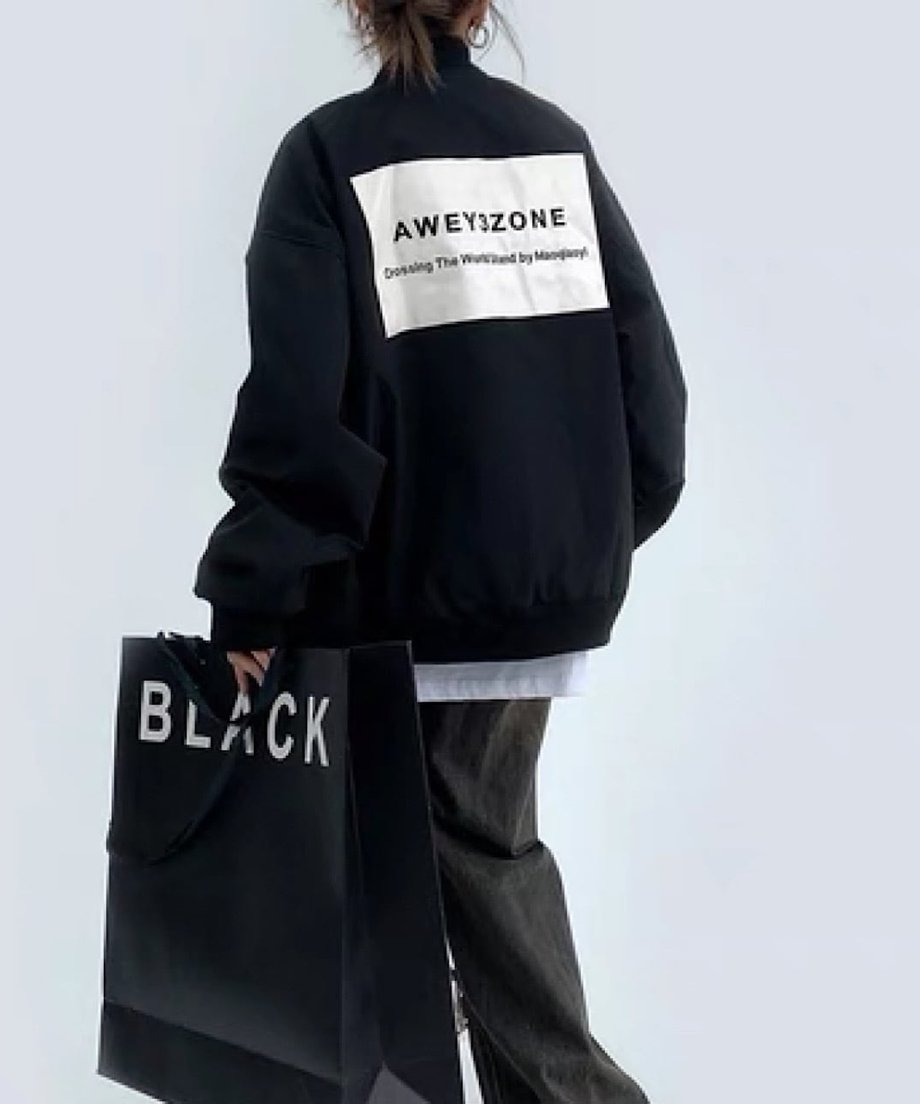 ブラックのオーバーサイズボンバージャケットを着用した後ろ姿。背中に白い帯状のブロックにロゴがプリントされたデザイン。黒い大きな紙袋を持っている。