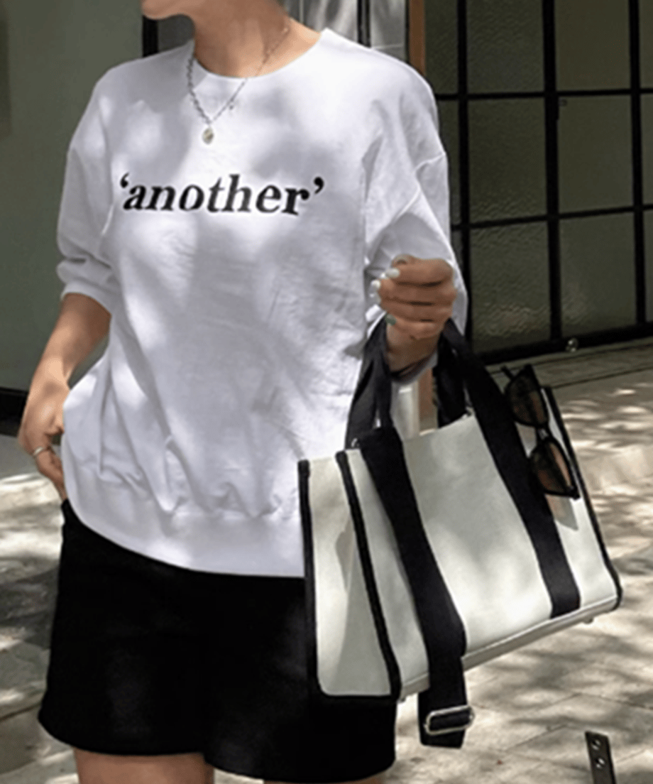 白地に「another」とロゴプリントされた五分袖のプルオーバーを黒のショートパンツに合わせ、白黒のキャンバストートバッグを持った女性のコーディネート。