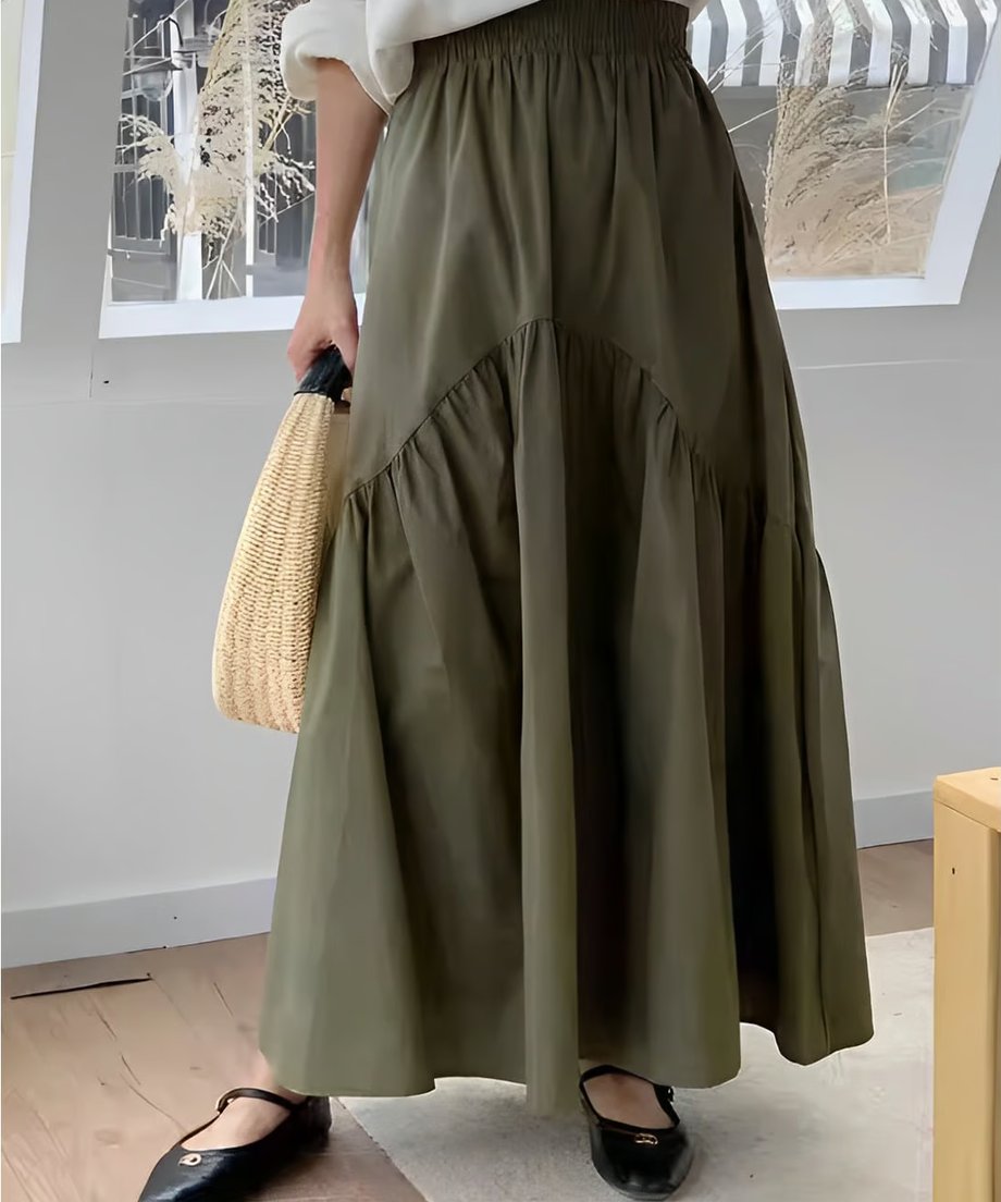 カーキ色のティアード切り替えマキシスカートを履いた女性のアップ。白いトップスをインし、麦わら素材のバッグを持っている。