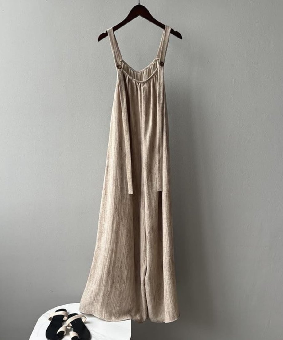 濃い茶色の木製ハンガーにかけられた、ベージュのワイドオーバーオールの胸元クローズアップ。肩紐のシンプルなデザインと、胸元のギャザーディテールがわかる。