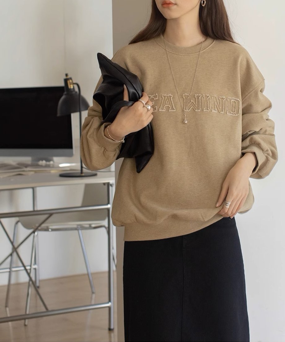 ベージュのロゴスウェットシャツを着た女性がオフィスのような場所で黒いバッグを持っている。