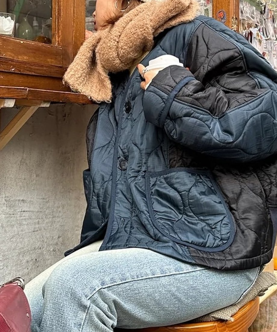 ネイビーブルーのツートンキルトジャケットを着用し、ベージュのマフラーを巻いた女性が椅子に座っている。ジャケットの大きなパッチポケット。
