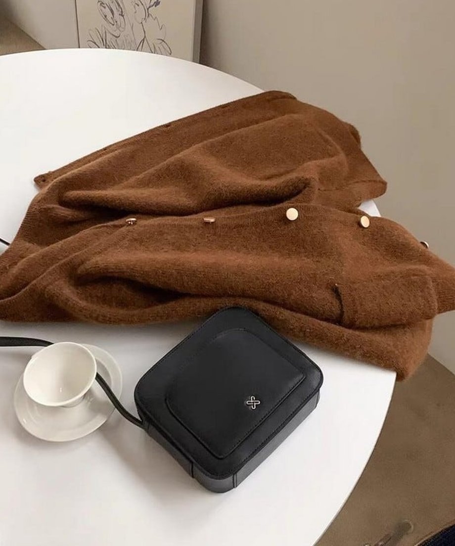 テーブルの上に、コーヒーのランタンスリーブカーディガンが畳まれており、黒いスクエア型のバッグと白いカップが並んでいる静物写真。