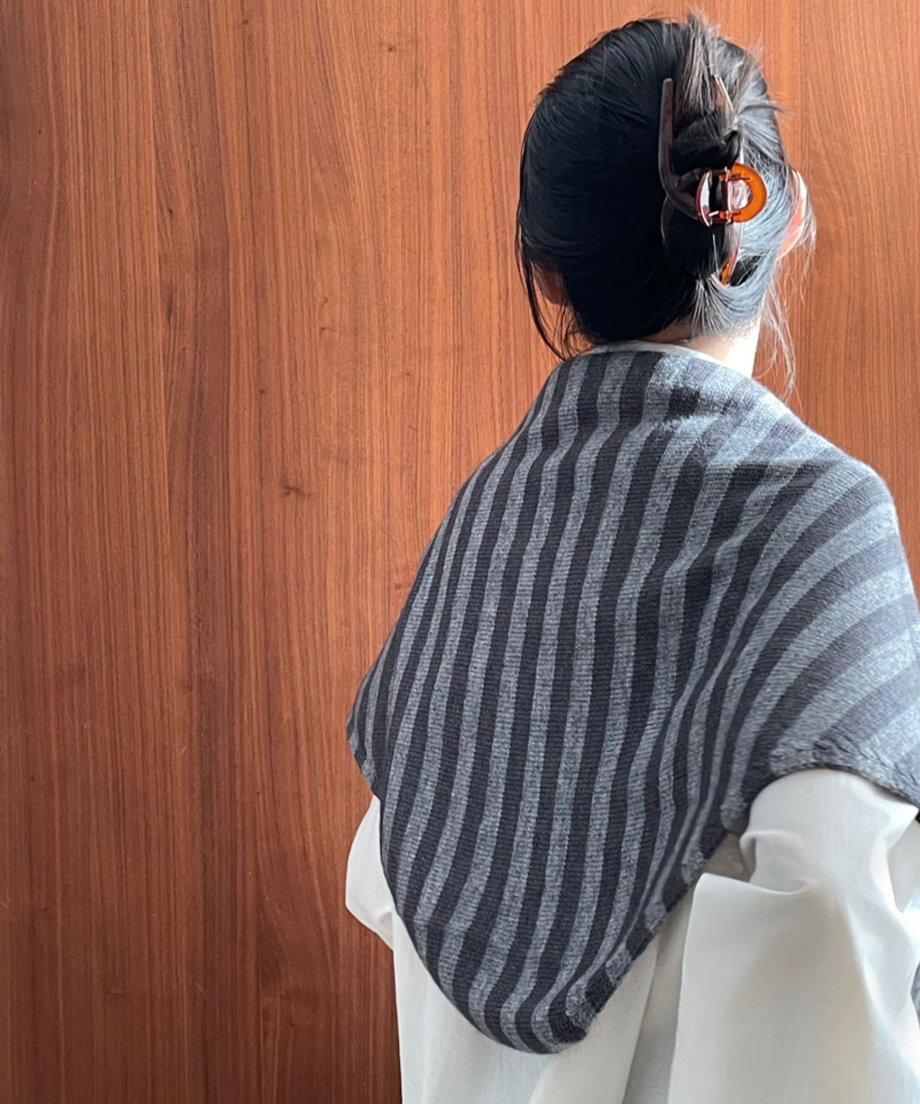 グレーストライプ柄のニットスカーフを着用した女性の後ろ姿。木製の壁の前で、髪をクリップでまとめている。